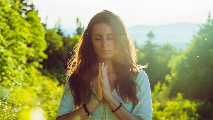 A woman standing outdoors with her hands at her heart, eyes closed